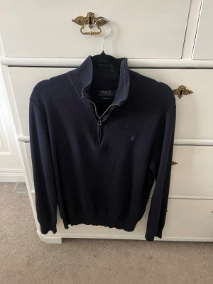 Mörkblå stickad tröja Polo Ralph Lauren - Mörkblå stickad tröja från Polo Ralph Lauren med halv dragkedja och hög krage. Tröjan har lång ärm, ribbade muddar och diskret broderad logga på bröstet. Perfekt för lager på lager och klassisk stil.