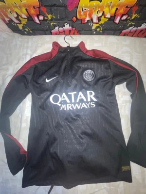 Svart PSG träningströja från Nike - Svart långärmad träningströja från Nike med PSG-logga och Qatar Airways-tryck på bröstet. Tröjan har röda detaljer längs ärmarna och halv dragkedja framtill. Perfekt för fotbollsträning eller sportiga dagar.