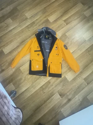 Gul vindjacka med tryck och huva - Snygg gul vindjacka med svarta detaljer och flera tryck på bröstet och ärmen. Jackan har huva, dragkedja och stora fickor framtill. Materialet är troligtvis polyester och jackan har en sportig look med patch på ärmen.