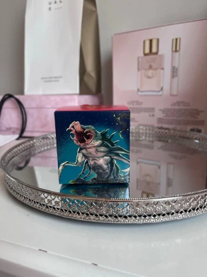 Stranger Things Tales from 85 box - Säljer en liten box med Stranger Things-tema, 'Tales from 85'. Boxen har färgstark design med motiv av en monsterliknande varelse och loggan i retrostil. Perfekt för fans av serien eller som samlarobjekt. Materialet är kartong och färgerna är mestadels blå, rosa och gul.
