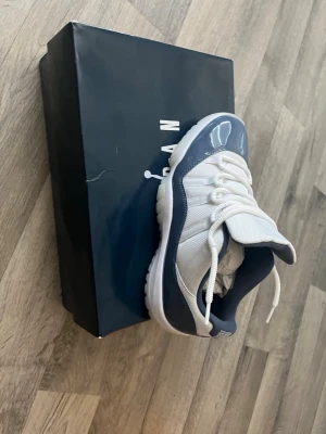 blåa jordan 11 - Snygga höga sneakers i vitt och marinblått med glansiga detaljer och klassisk snörning. Ovandelen är i textil och syntet, med en vit sula som ger en sportig look. Perfekta för dig som vill sticka ut med stilrena färger och modern design.
