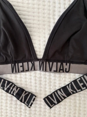 Svart bikiniöverdel Calvin Klein - Observera ! Trasig band på ryggen. Det går att knyta dock.Snygg svart bikiniöverdel från Calvin Klein med bred resår och logga runt om.  Tillverkad i mjukt material som sitter bekvämt.