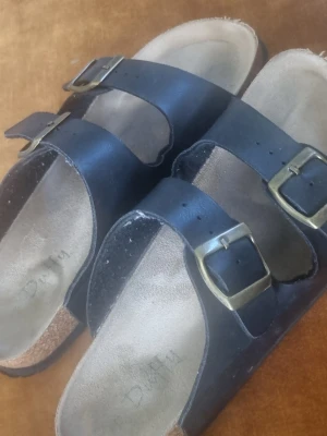 Svarta sandaler  - Säljer ett par svarta sandaler med två breda remmar och justerbara metallspännen. Fotbädden är i mockaliknande material och sulan har ett vågigt mönster för bra grepp. Klassisk slip-in modell som passar till många stilar.