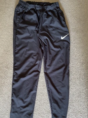 Svarta träningsbyxor från Nike - Säljer ett par svarta träningsbyxor från Nike med klassisk vit swoosh-logga på benet. Byxorna har resår i midjan och dragsko för bästa passform samt sidofickor. Perfekta för gymmet eller löprundan.