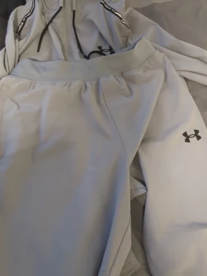  Under Armour tracksuit/set/träningsset - Säljer ett grått träningsset från Under Armour med byxor och zip-hoodie. Setet har svarta loggor och detaljer, perforerade partier för ventilation och dragsko i huvan. Perfekt för träning eller chill. Materialet är lätt och funktionsanpassat. Jätte bra skick storlek m den har en fläck där könsorganet ligger av mat men de går o ta bort men syns inte så mkt bara när de jätte ljust