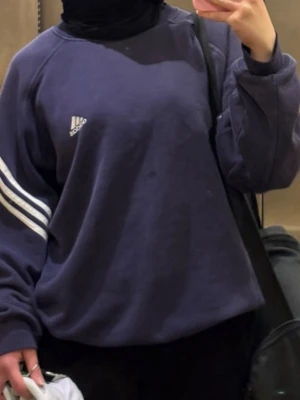Mörkblå Adidas stickad tröja - Säljer en mörkblå stickad tröja från Adidas med klassiska vita ränder på ena ärmen och Adidas-logga på bröstet. Tröjan har en avslappnad passform och långa ärmar, perfekt för en sportig och chill look.