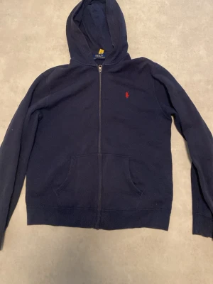  Äkta köpt på NK! Mörkblå hoodie från Polo Ralph Lauren - Mörkblå hoodie med dragkedja från Polo Ralph Lauren. Klassisk design med röd broderad logga på bröstet, huva och kängurufickor framtill. Tillverkad i mjukt bomullsmaterial som är skönt att bära. Perfekt för en avslappnad och stilren look. NYPRIS 1200kr