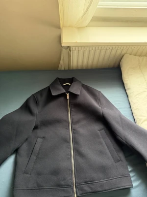 Svart jacka med dragkedja - Stilren svart jacka med rak passform och klassisk krage. Jackan har hel dragkedja framtill och två snedställda fickor. Materialet ser ut att vara ullblandning, perfekt för kyligare dagar. Enkel och tidlös design som passar till det mesta.
