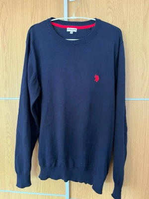 Mörkblå tröja från U.S. Polo Assn - Klassisk mörkblå tröja från U.S. Polo Assn med rund halsringning och röd broderad logga på bröstet. Tröjan har ribbade muddar vid ärmslut och nederkant. Perfekt för en stilren och avslappnad look.