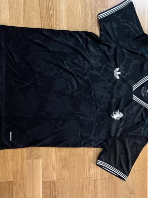 Svart Adidas t-shirt med blommönster - Snygg svart t-shirt från Adidas med diskret blommönster i tyget. Klassisk Adidas-logga på bröstet och vita ränder på krage och ärmslut. Tillverkad i polyester för en sportig känsla. Perfekt för dig som gillar streetwear och vill sticka ut.    Fraktar samma eler dagen efter du köpt!!! Endast en kvar i lager!!!