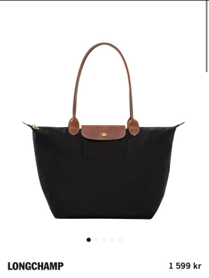 Svart handväska från Longchamp - Klassisk svart handväska från Longchamp med bruna läderhandtag och lock. Väskan har en enkel och stilren design med guldfärgad knappdetalj och dragkedja. Perfekt för dig som gillar tidlös och snygg stil.
