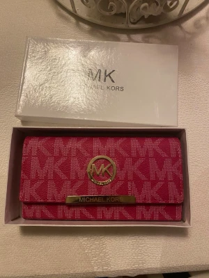 Rosa plånbok/handväska från Michael Kors - Snygg rosa plånbok från Michael Kors med MK-logga i guld framtill och mönstrat tyg med MK-bokstäver. Plånboken har flera fack inuti och stängs med tryckknapp. Perfekt för dig som vill ha en trendig accessoar med plats för kort och kontanter.