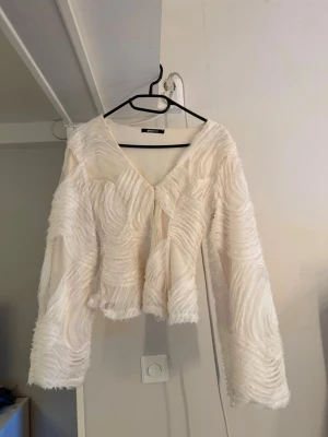 Vit blus med struktur och vida ärmar - Säljer en unik vit blus med v-ringning och vida, långa ärmar. Blusen har ett strukturerat mönster med vågiga detaljer som ger ett coolt och trendigt intryck. Perfekt för dig som vill sticka ut med en statement topp.
