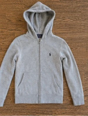 Grå zip hoodie med huva - Snygg grå hoodie med dragkedja och huva. Klassisk design med två fickor framtill och en liten broderad logga på bröstet. Tillverkad i mjukt sweatshirtmaterial som är perfekt för chill dagar. Långärmad och ribbade muddar vid ärmslut och nederkant.