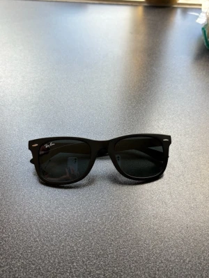 Svarta Ray-Ban Wayfarer solglasögon - Klassiska svarta Wayfarer solglasögon från Ray-Ban med mattsvart båge och mörka glas. Snygg och tidlös design med Ray-Ban-logga på linsen och silverdetaljer vid fronten. Perfekta för soliga dagar och ger en cool vibe till din outfit.