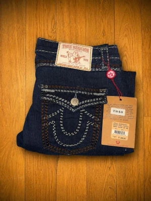 True Religion Ricky Super QT Jeans – Made in USA – Nya med prislapp – W31 - Helt oanvända True Religion Ricky Super QT-jeans med originalprislappar kvar.  • Storlek: W31 (M) • Made in USA • 100 % bomull • Nyskick – aldrig använda  Mått (mätta plant): • Midja: 40 cm • Innerbenslängd: 76 cm • Benlängd fram: 97 cm • Benlängd bak: 102 cm