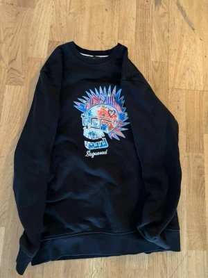 Svart sweatshirt med färgglad dödskalle - Cool svart sweatshirt med ett stort färgglatt dödskalletryck på bröstet och texten 'Dsquared' under motivet. Tröjan har rund halsringning och långa ärmar. Perfekt för dig som gillar streetwear och unika prints. Pris kan diskuteras vid snabb affär!