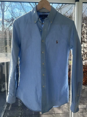 Ljusblå skjorta från Ralph Lauren - Klassisk ljusblå skjorta från Ralph Lauren med button-down krage och broderad logga på bröstet. Skjortan har långa ärmar, knappar framtill och är tillverkad i mjuk bomull. Perfekt för en clean och stilren look.