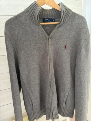  Polo Ralph Lauren grå zip - Snygg grå ziptröja från Polo Ralph Lauren med ribbad krage och muddar. Tröjan har dragkedja framtill, två fickor och den klassiska broderade loggan på bröstet. Perfekt till lager på lager och enkel att matcha med olika outfits.