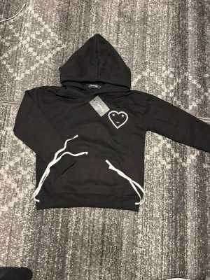 Svart hoodie med broderat hjärta - Svart hoodie med huva och vita dragsnören. På bröstet finns ett broderat hjärta med ett ansikte i vitt, vilket ger en unik och trendig look. Hoodien har en klassisk passform och är tillverkad i mjukt material som känns skönt mot huden.