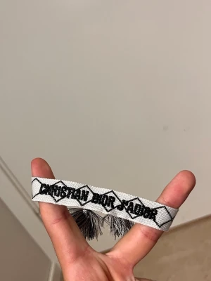 Vitt tygarmband från Dior - Säljer ett vitt tygarmband från Christian Dior med svart broderad text och grafiskt mönster. Armbandet har tofsar i ändarna och är justerbart för att passa olika handleder. Perfekt accessoar för dig som gillar exklusiva detaljer.