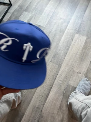 trapstar Blå snapback keps med vit brodyr - Snygg blå snapback keps med stor vit brodyrtext runt hela kepsen. Klassisk platt skärm och justerbar baksida med plastspänne. Passar perfekt för dig som vill sticka ut med en streetwear-look.