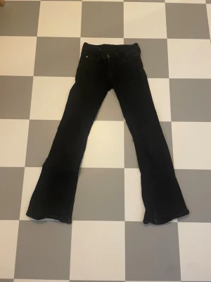 Svarta bootcut jeans 25/30 - Säljer ett par svarta bootcut jeans med klassisk femficksmodell och utsvängda ben. Jeansen har knapp- och dragkedjestängning samt en lila patch bak i midjan. 