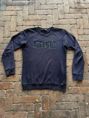 Mörkblå sweatshirt med Fendi-tryck - Mörkblå sweatshirt med rund hals och långa ärmar. Framsidan har ett svart Fendi-tryck i blockbokstäver. Tröjan har ribbade muddar vid ärmslut och nederkant, perfekt för en avslappnad och trendig look.