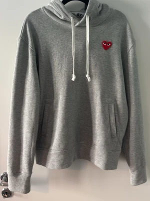 Cdg hoodie  - Säljer en grå hoodie från Comme des Garçons Play med den ikoniska röda hjärtloggan på bröstet. Hoodien har huva med vita snören och två fickor framtill. Tillverkad i mjukt bomullsmaterial, perfekt för en chill och stilren look.