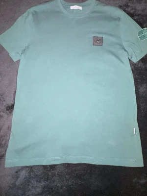 Grön t-shirt från Stone Island - Snygg grön t-shirt från Stone Island med klassisk patch på ärmen och diskret logga på bröstet. Tillverkad i mjuk bomull och har en normal passform med korta ärmar. Perfekt till jeans eller shorts för en clean look.