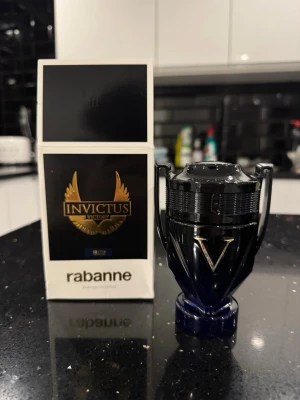 Paco Rabanne Invictus Victory elixir - Parfymflaska från Paco Rabanne, modell Invictus Victory. Flaskan har en unik pokalform i svart och mörkblått med silverdetaljer och ett stort V på framsidan. Förpackningen är svart och vit med guldiga detaljer och logga. En riktigt snygg och modern doftaccessoar.