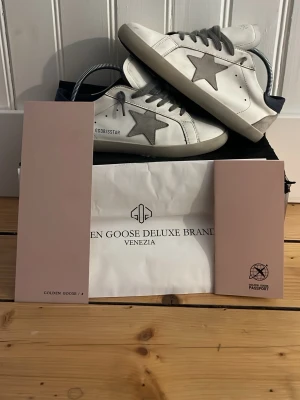 Golden Goose vita sneakers med grå stjärna - Golden Goose sneakers i vitt läder med grå mockadetaljer och ikonisk stjärna på sidan. Skorna har grå snörning, svart hälparti och klassisk låg siluett. Kommer med originalpåse och förpackning. Perfekta för en trendig och avslappnad stil. Utan kvitto och köpt på Cn-Fans
