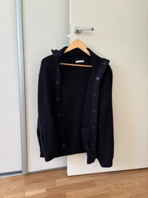 Mörkblå stickad kofta från ZARA - Säljer en mörkblå, grovstickad kofta från ZARA med knappar framtill och hög krage. Koftan har långa ärmar och ribbstickad struktur, perfekt för lager på lager. Klassisk och enkel design som passar till det mesta.