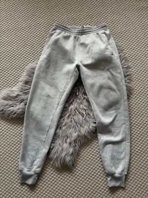 Gråa mjukisbyxor med resår - Säljer ett par klassiska gråa mjukisbyxor med resår i midjan och vid bensluten. Byxorna har en avslappnad passform och är tillverkade i mjukt bomullsmaterial. Perfekta för chill dagar hemma eller när du vill vara bekväm.