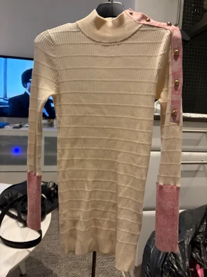  Tröja storlek xs - Ribbstickad tröja i beige med kontrasterande rosa muddar och axelparti. Hög hals och dekorativa guldknappar längs ena axeln ger en unik look. Långärmad och figurnära modell, perfekt för dig som gillar stilrena plagg med en twist.