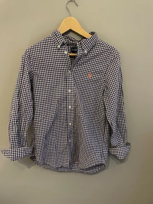 Rutig skjorta från Ralph Lauren - Klassisk blå och vit rutig skjorta från Ralph Lauren med orange broderad logga på bröstet. Skjortan har lång ärm, knappar framtill och button-down krage. Perfekt för en stilren och tidlös look.