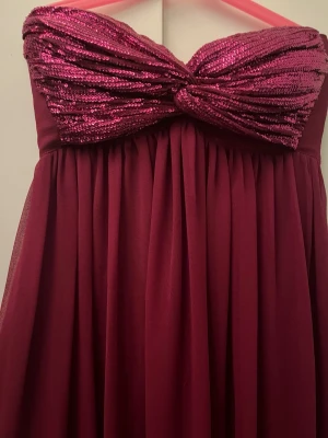 Vinröd långklänning med paljetter - Snygg vinröd långklänning med axelbandslös design och glittrig paljettbyst. Priset går att diskuteras🥰Klänningen har ett svepande fall och markerad byst med twistdetalj. Perfekt för dig som vill sticka ut på bal eller fest.