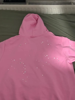 Rosa hoodie från Sp5der med stjärnor - Säljer en rosa hoodie från Sp5der i storlek L. Hoodien har en huva och är dekorerad med små vita och silvriga stjärnor på ryggen. Tillverkad i 100% bomull och har en cool streetwear-vibe. Perfekt för dig som gillar unika detaljer och vill sticka ut.