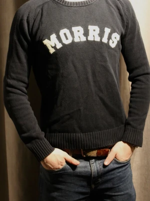 Svart stickad tröja från Morris - Snygg svart stickad tröja från Morris med vit broderad logga över bröstet. Tröjan har rund halsringning och ribbade muddar vid ärmslut och nederkant. Perfekt för en avslappnad och stilren look.