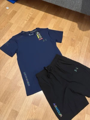 Under Armour träningsset blå/svart storlek M - Snyggt träningsset från Under Armour med en mörkblå t-shirt och svarta shorts. T-shirten har kort ärm och logga samt text i regnbågsfärger. Shortsen har elastisk midja med dragsko och matchande logga. Perfekt för gym eller löpning.