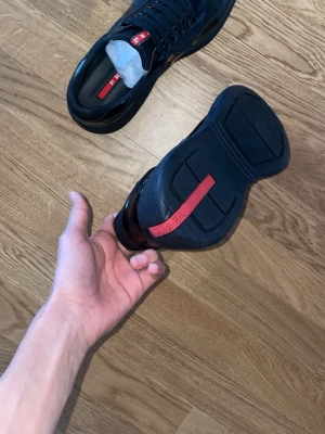Svarta cups  - Snygga svarta sneakers från Zara med glansiga och mesh-detaljer. Skorna har en röd logotyp på plösen och en röd rand under sulan. Klassisk låg modell med snörning och chunky sula. Perfekta för dig som vill ha en stilren och exklusiv look. Skorna har aldrig varit använda.