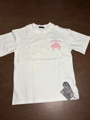 Vit t-shirt Chrome Hearts Caution - Vit t-shirt från Chrome Hearts med rosa tryck fram och bak. Framsidan har ett litet Chrome Hearts-logotyptryck i rosa, medan baksidan har ett stort 'Caution'-tryck och grafiska detaljer i rosa och svart. Klassisk passform och rund hals. Perfekt för dig som gillar streetwear.