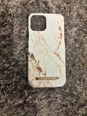 Vitt marmormönstrat mobilskal iDeal iPhone 12 - Snyggt mobilskal från iDeal of Sweden med vitt marmormönster och inslag av guldiga och bruna ådror. Skalets material är hårdplast och har en guldfärgad logga längst ner. Passar perfekt för dig som vill ha en stilren och trendig look till mobilen. Pris kan diskuteras 