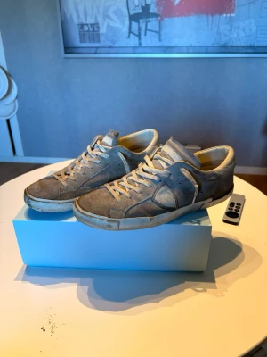 Grå Philippe Model sneakers i mocka - Snygga grå sneakers från Philippe Model i mjuk mocka med vita detaljer och vita skosnören. Klassisk låg modell med gummisula och diskret logga på sidan. Perfekta för dig som gillar stilrena och trendiga skor.