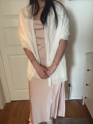 Ljusbeige långklänning i satin - Elegant ljusbeige långklänning i satin med slits på sidan. Klänningen har en figurnära passform och är ärmlös med enkel, stilren design. Perfekt för dig som gillar en klassisk och sofistikerad look.