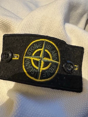 Vit stickad tröja Stone Island M - Säljer en vit stickad tröja från Stone Island i storlek XXL. Tröjan har klassisk svart och gul Stone Island patch på ärmen och ribbstickad krage med svarta och vita ränder. Perfekt för dig som gillar streetwear och exklusiva märken.