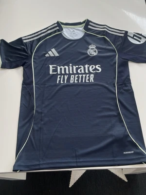 Real Madrid tränings t-shirt Adidas - Mörkblå Real Madrid tränings t-shirt från Adidas med vita detaljer och klubbmärke på bröstet. Tillverkad i funktionsmaterialet Aeroready som andas. Klassiska Adidas-ränder på axlarna och tryck med 'Emirates Fly Better' på framsidan.