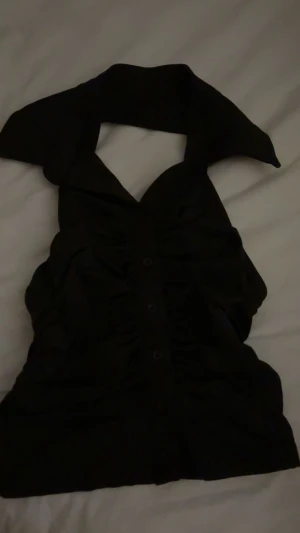 Svart halterneck topp med knappar - Snygg svart halterneck topp med bred krage och knappar framtill. Toppen har en figurnära passform och rynkade detaljer längs sidorna. Perfekt för dig som vill ha en trendig och stilren look.