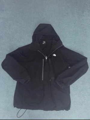Svart vindjacka från The North Face - Svart vindjacka från The North Face med huva och dragkedja framtill. Jackan har två vertikala fickor med dragkedja och justerbar nederkant. Perfekt för blåsiga dagar och enkel att matcha med olika outfits.