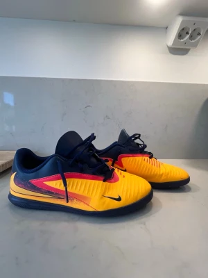 Nike gula och blå fotbollsskor - Säljer ett par Nike fotbollsskor för inomhusbruk i en snygg gul färg med blå och röda detaljer. Skorna har snörning, låg profil och slitstark sula i gummi för bra grepp på inomhusplan. Perfekta för dig som vill sticka ut på planen!